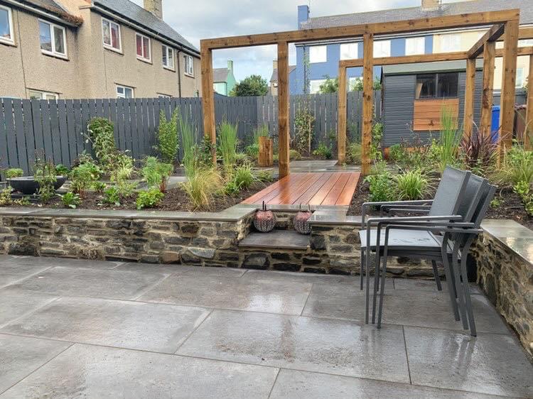 Grey patio with a stone retaining wall, timber pergola and new planting